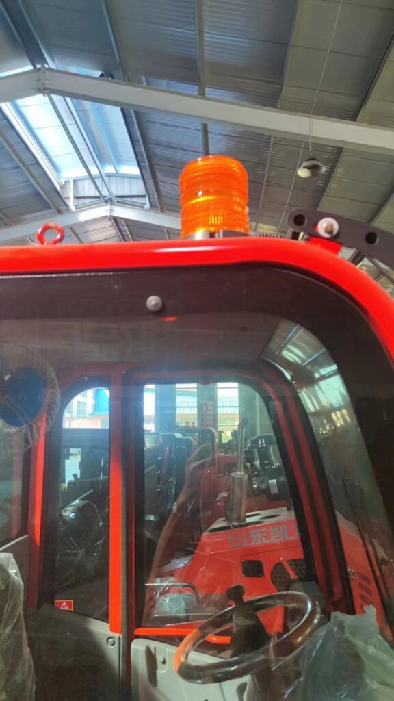 A photo of a yellow and black four-wheel drive forklift with a flashing warning light on top of its mast in a factory setting.