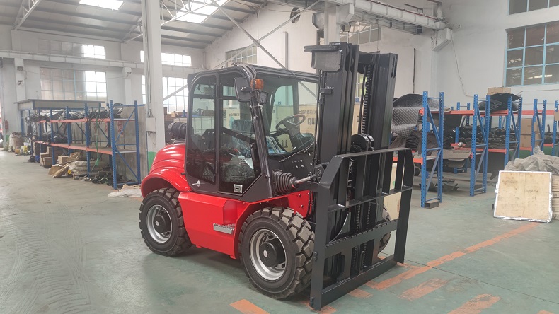 A customized off-road forklift with a red body, a black cab, a black mast, and a green load on the forks. The forklift is driving on a dirt road in a forest. The logo of Kaystar Heavy Industries is visible on the side of the cab. 