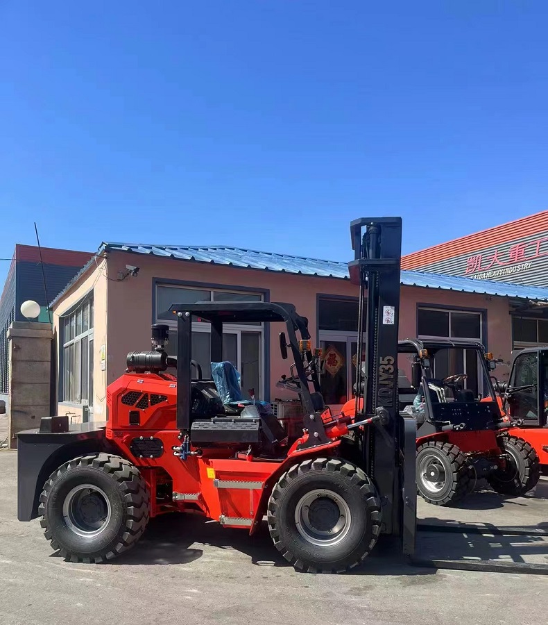 A photo of a red and black forklift with four large wheels, a rear-mounted articulated joint, a simple cab, and a three-stage mast with forks. The forklift is parked on a dirt road with trees and hills in the background. The logo of Kaystar is visible on the side of the cab.