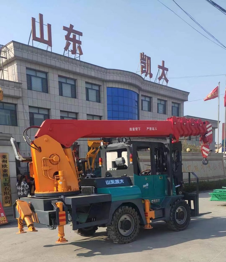 A photo of a blue Pioneer50 forklift that has been modified into an off-road mini crane with a red boom and four yellow support legs. The mini crane is lifting a large blue container on a green field with the kaystar factory  in the background.