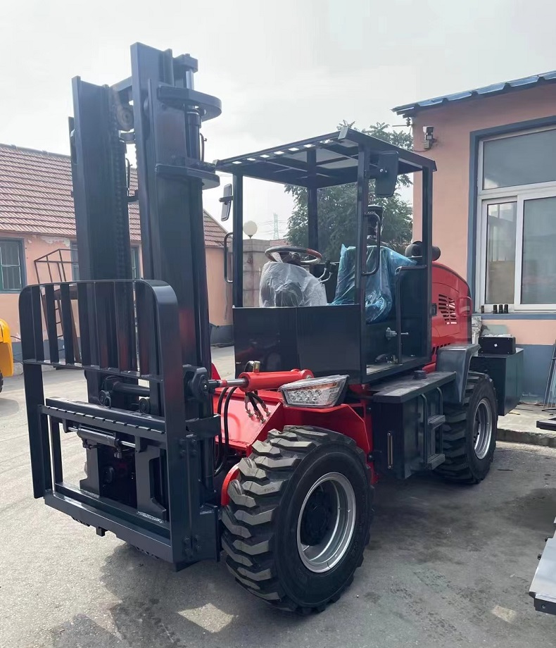 A photo of a red articulated four-wheel drive mini forklift with a rated load of 3 tons and a body width of 1.5 meters. The forklift is parked on a dirt road surrounded by trees and plants.（left side view）