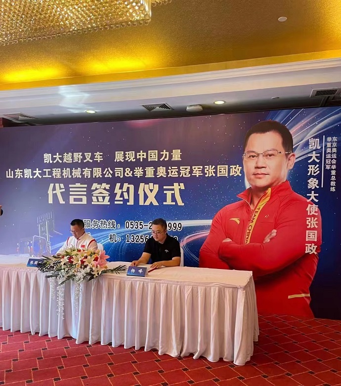 A picture of two men shaking hands while holding a contract paper. One man is wearing a suit and tie, while the other man is wearing a sports jacket and a medal. Behind them are several banners with the logo of KAIDA and the words “Four-Wheel Forklift”.
