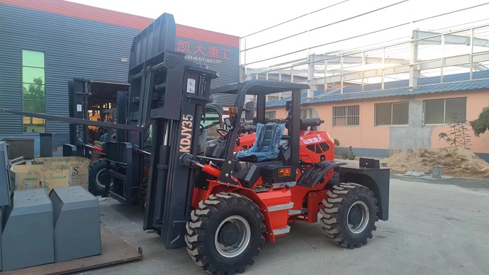 A redand black forklift with four large wheels and a long mast with two forks at the end. The forklift has a rear-articulated design and a detachable simple driving shed. The forklift is parked on a dirt road with some trees and buildings in the background.